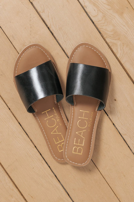 Coconuts by Matisse Cabana Slides in black, featuring wide straps, displayed on a light wooden floor.