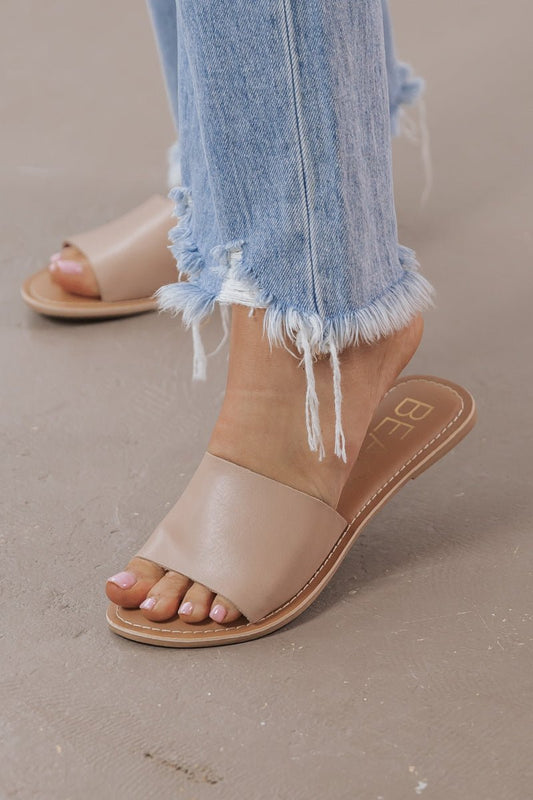 A person in light-wash, frayed-hem jeans wears Coconuts by Matisse Cabana Slides - Natural on a neutral surface.