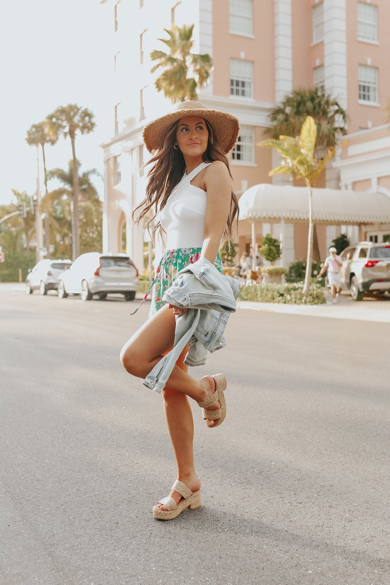 Woman in sun hat and summer outfit wears the Cross Front Halter Ribbed Bodysuit - FINAL SALE, with palm trees and pink building behind.