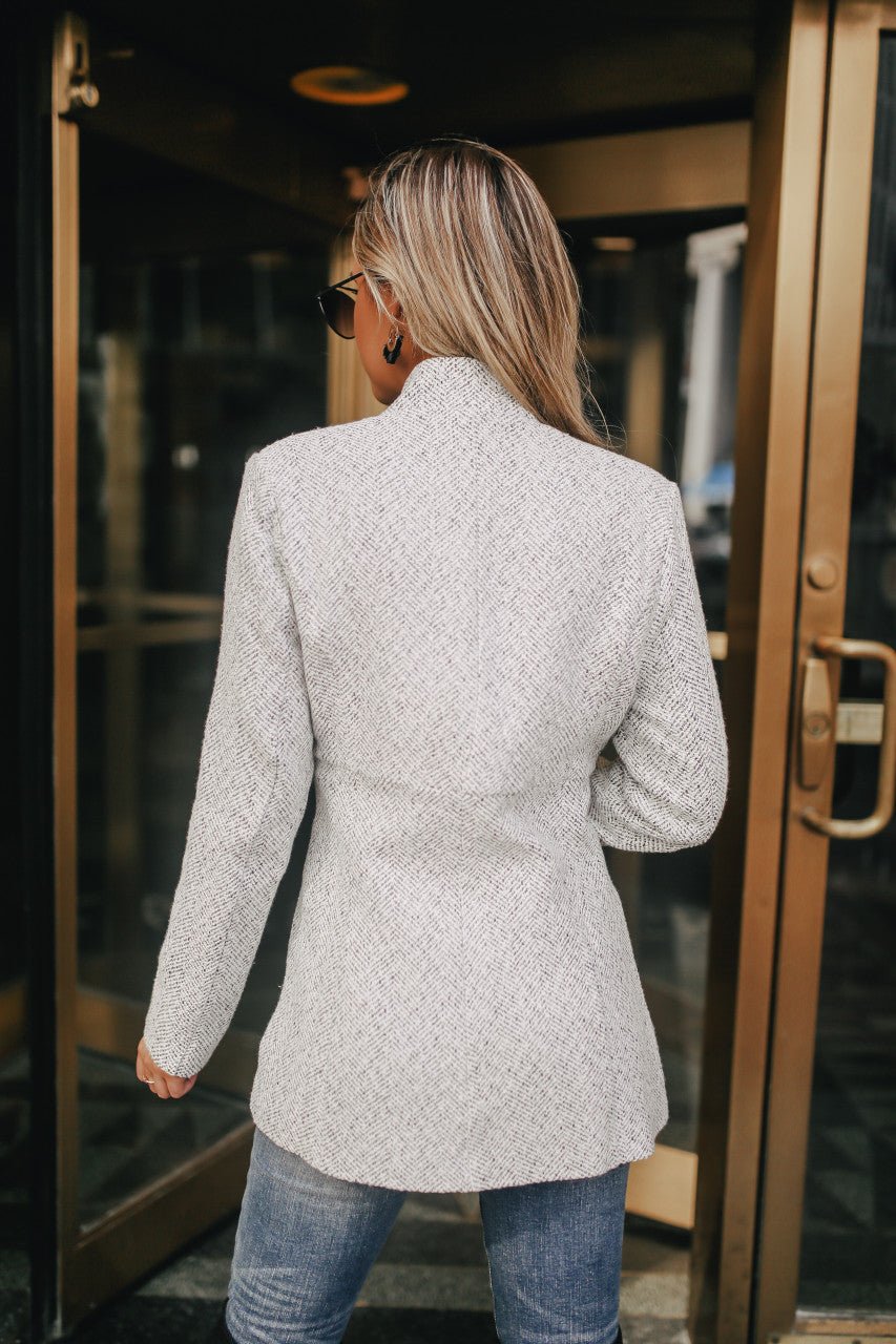 A woman with long blonde hair wears the Open Front Herringbone Jacket - Grey/White and jeans, facing away before glass doors.