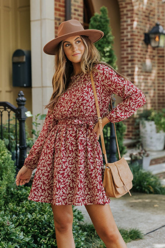 Woman wears the Sidney Maroon Floral Print Long Sleeve Dress, posing outdoors by a brick building and greenery.