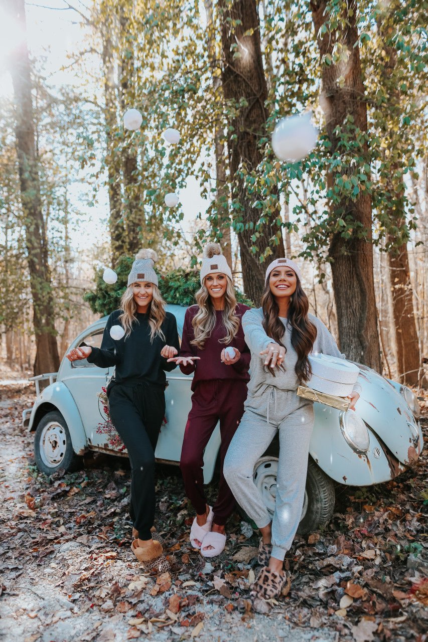 Three women in Step Up Lounge Sets and beanies have a playful snowball fight by a vintage car in a sunlit forest.