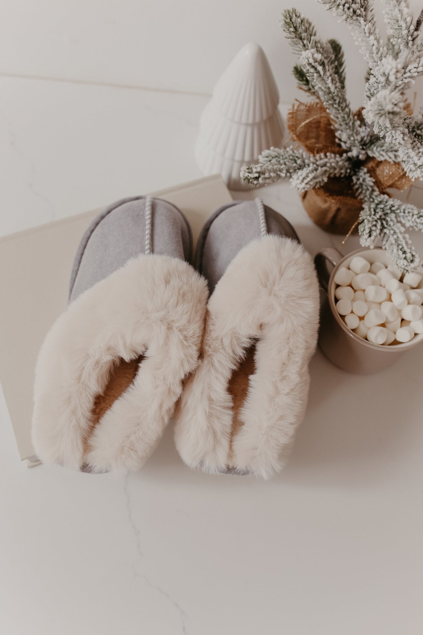 The Sweet Dreams Grey Faux Sheepskin Slippers rest beside a mug of marshmallows, a book, and snowy holiday décor.