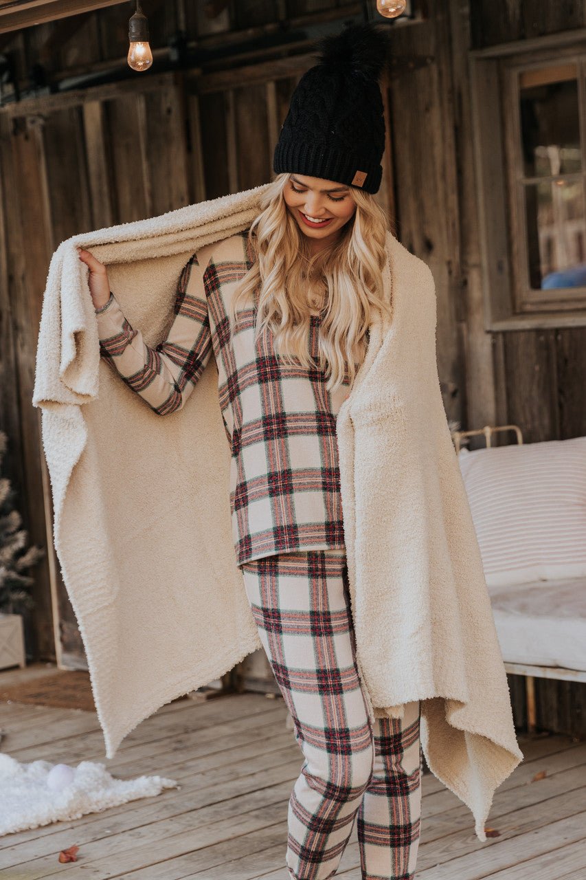 A woman in Taupe Tartan Plaid Jogger Pajama Pants and a black knit hat smiles on a porch, wrapped in a cozy beige blanket.