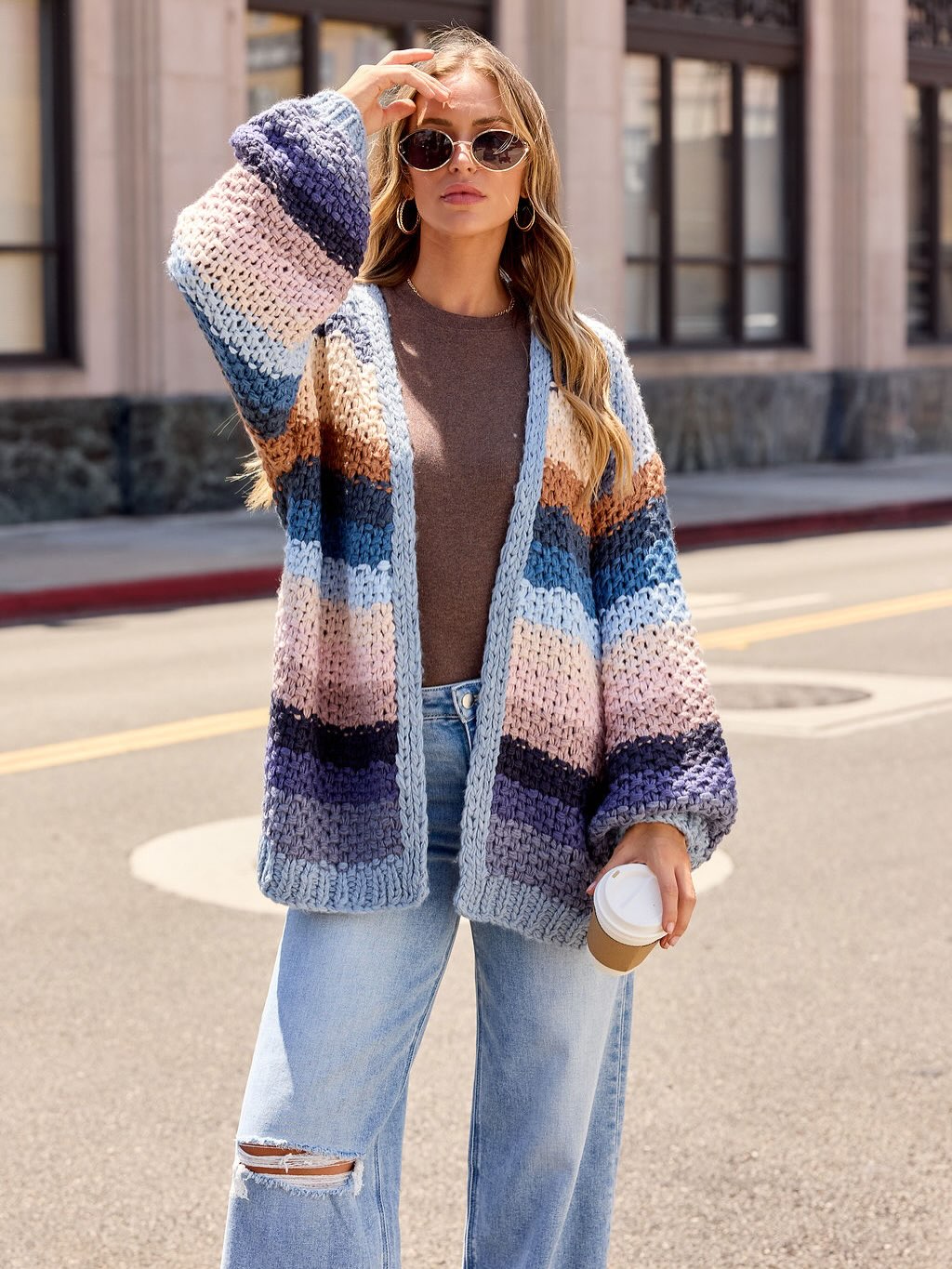 A woman wearing sunglasses, a brown shirt, and a colorful, striped knit cardigan stands on a city street holding a coffee cup, with one hand adjusting her sunglasses.