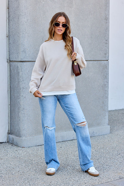 Woman in sunglasses and The Everyday Pullover - Clay, ripped jeans, and sneakers stands outside holding a brown handbag.