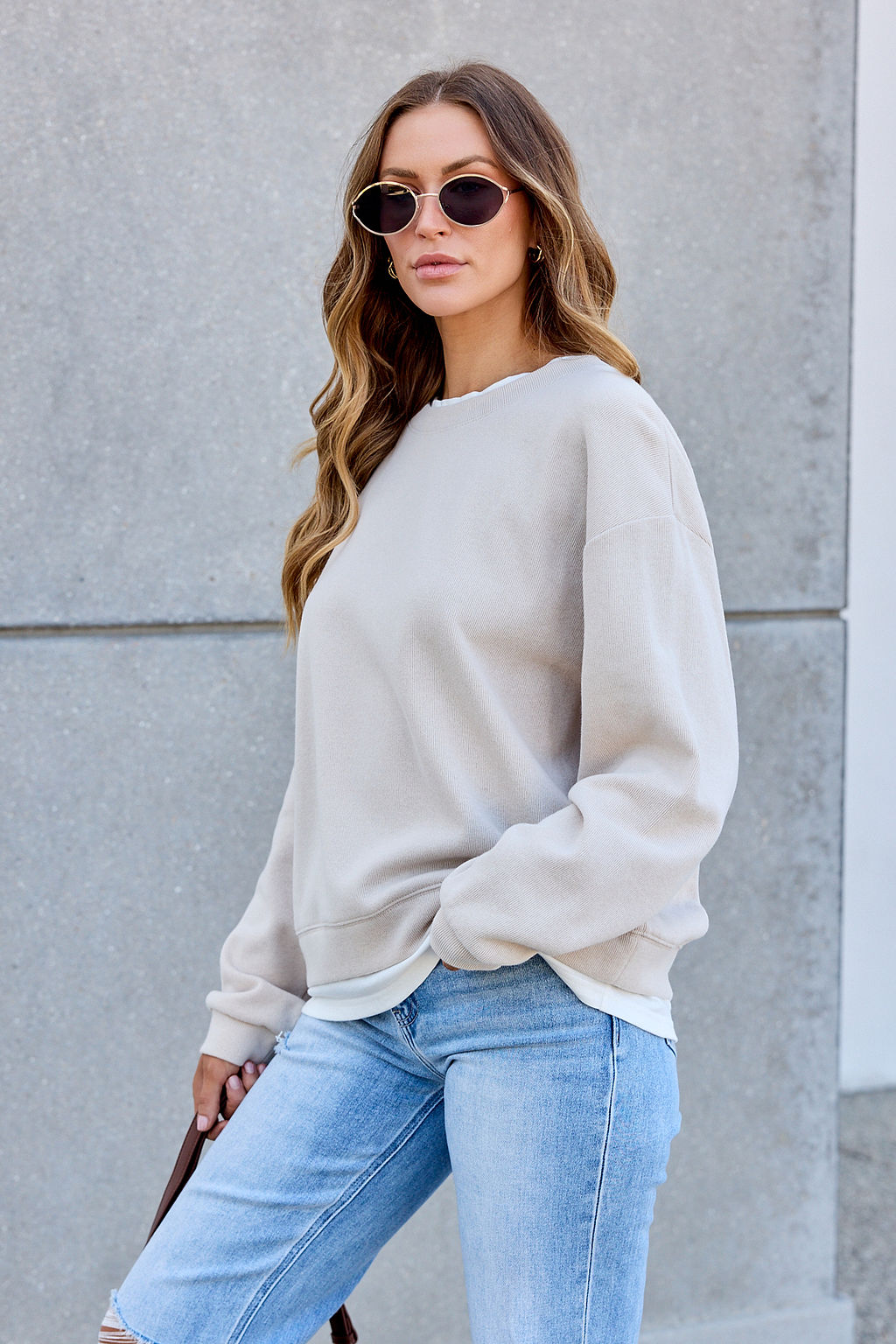 Woman in The Everyday Pullover - Clay and ripped jeans leans on a gray wall, radiating confidence and essential style.