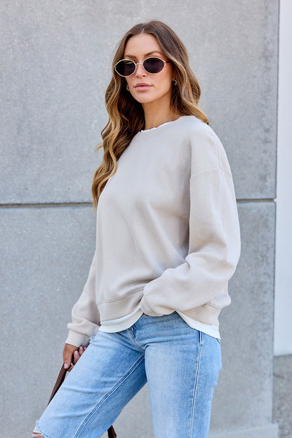Woman in The Everyday Pullover - Clay and ripped jeans leans on a gray wall, radiating confidence and essential style.