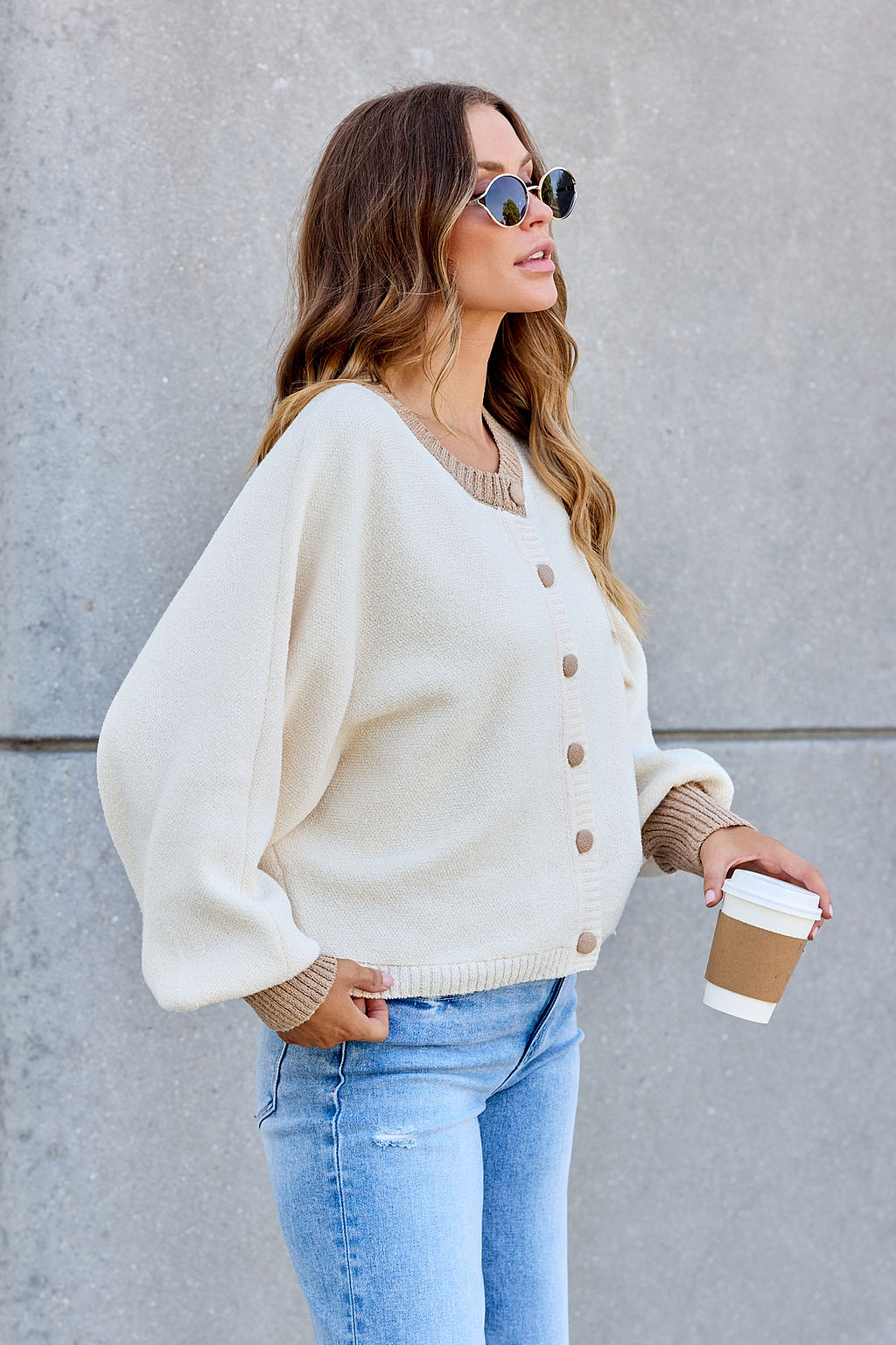 A woman in sunglasses wears the Contrast Ribbed Cardigan - Cream, holding a coffee cup by a gray concrete wall.