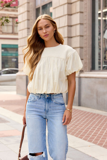 Woman with long wavy hair in the Bubble Hem Short Sleeve Top - Ivory stands on a city sidewalk, holding a brown purse.