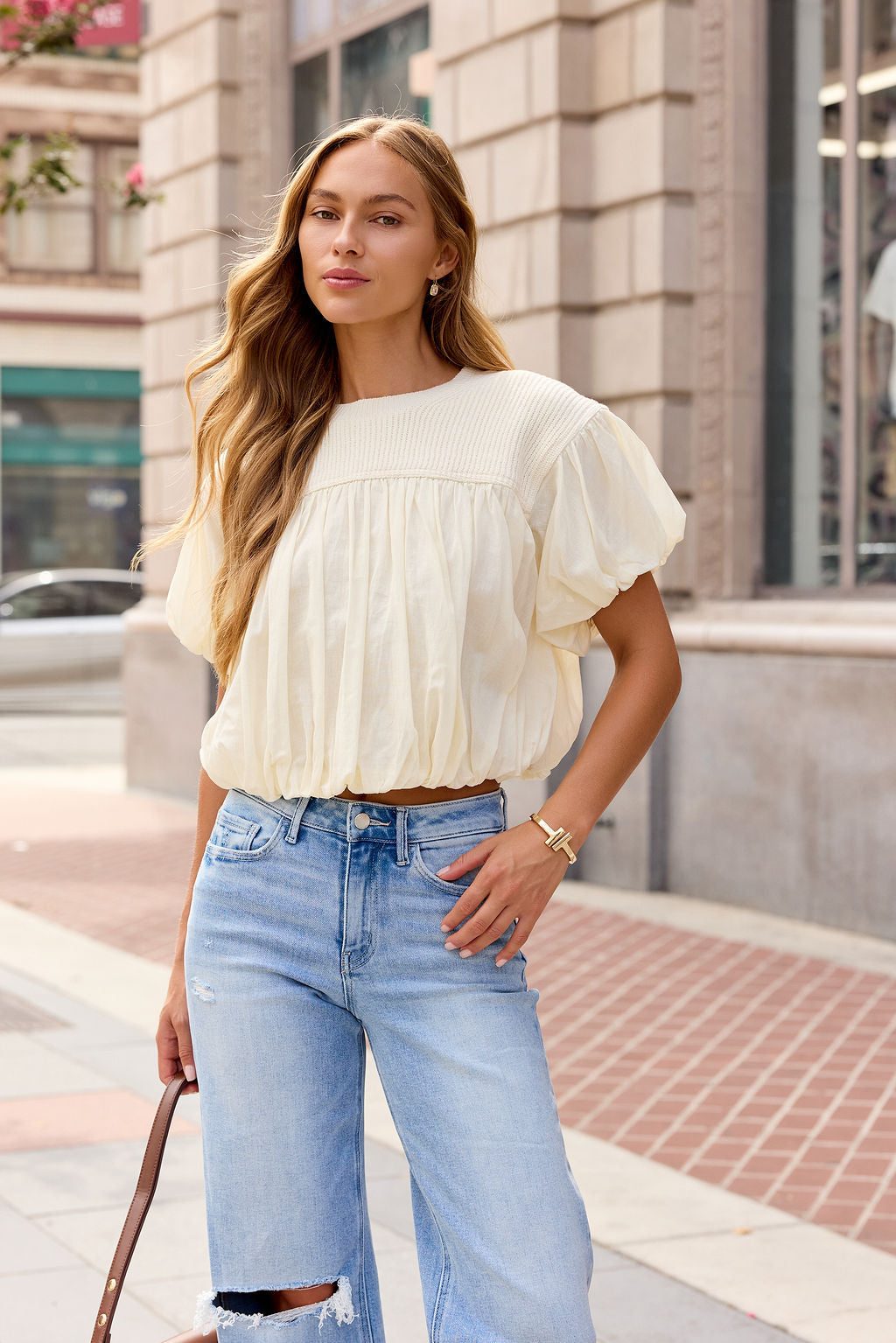 Woman in the Bubble Hem Short Sleeve Top - Ivory stands on a city sidewalk, holding a brown strap and wearing ripped jeans.
