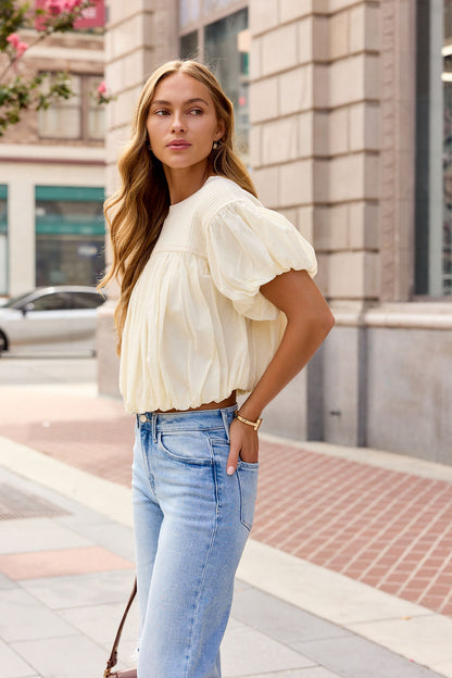 Woman in the Bubble Hem Short Sleeve Top - Ivory stands on a city sidewalk, looking to the side.