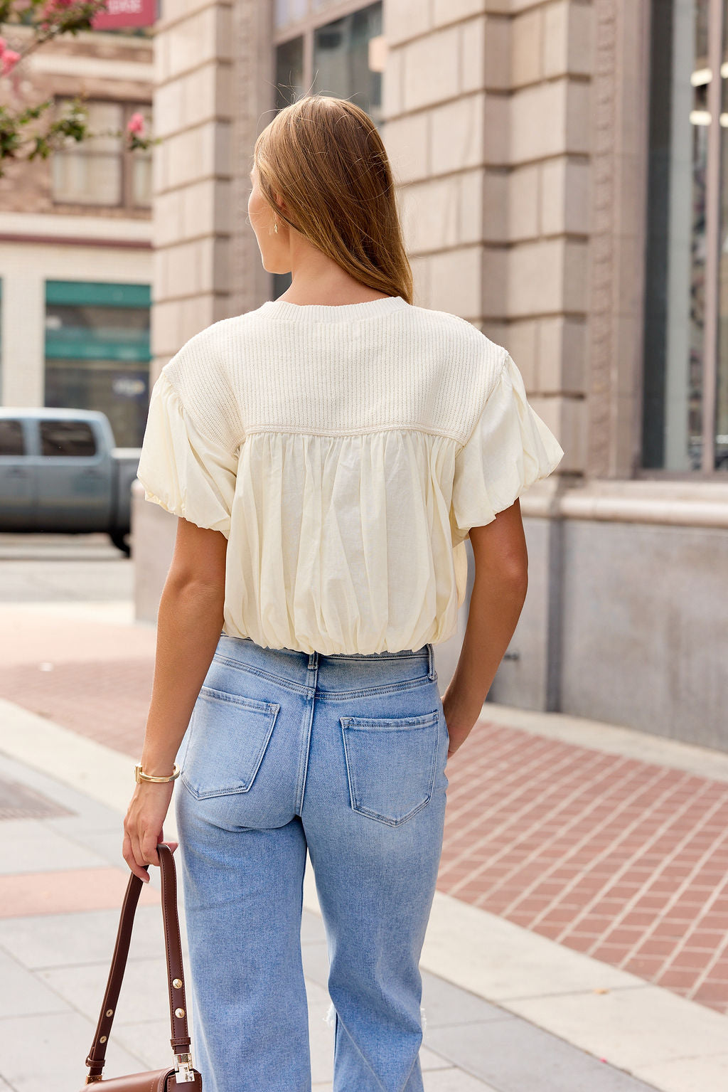 Woman with long hair wears the Bubble Hem Short Sleeve Top in Ivory with light blue jeans, standing on a city sidewalk, back to camera.
