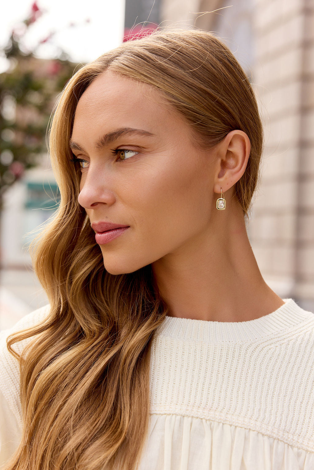 Woman with long, wavy blonde hair in a cream sweater wears Emerald Cut Drop Earrings - Gold while looking left outdoors.