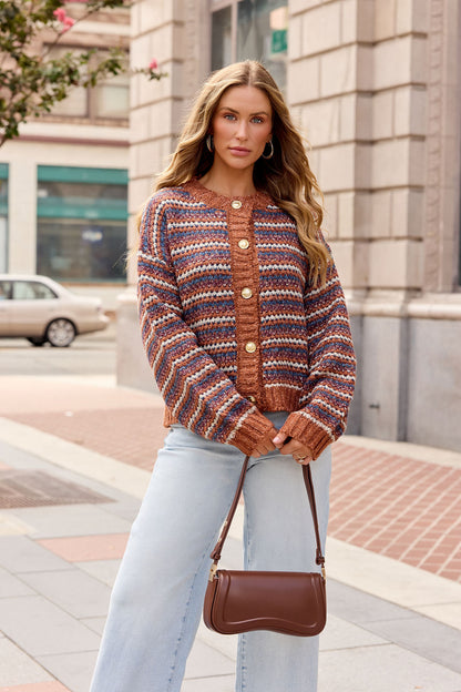 Woman in a Multi Striped Button Down Sweater and light jeans stands on city sidewalk, carrying a brown handbag.