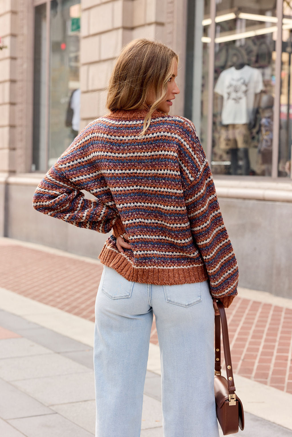 A woman in the Multi Striped Button Down Sweater and light jeans stands on a city sidewalk, her back to the camera.