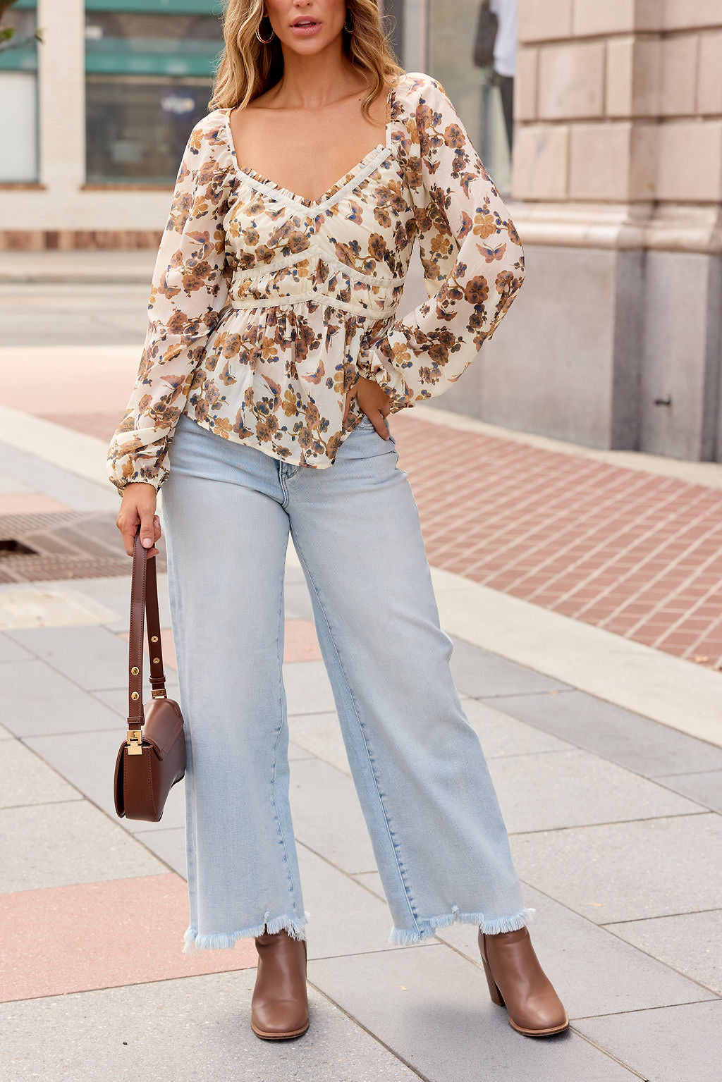 Woman in the Hazel Floral Print Top - Cream stands on a city sidewalk, styled with jeans, brown boots, and a matching handbag.