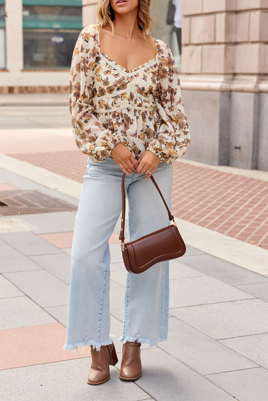 Woman in a Hazel Floral Print Top - Cream, light blue frayed jeans, brown boots, and handbag on a city sidewalk.