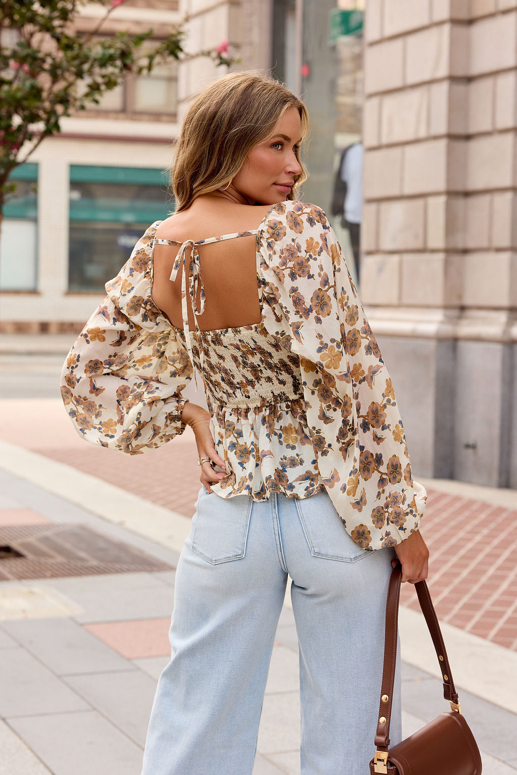 Woman with wavy hair wears the Hazel Floral Print Top - Cream, light jeans, and holds a brown handbag on a city sidewalk.