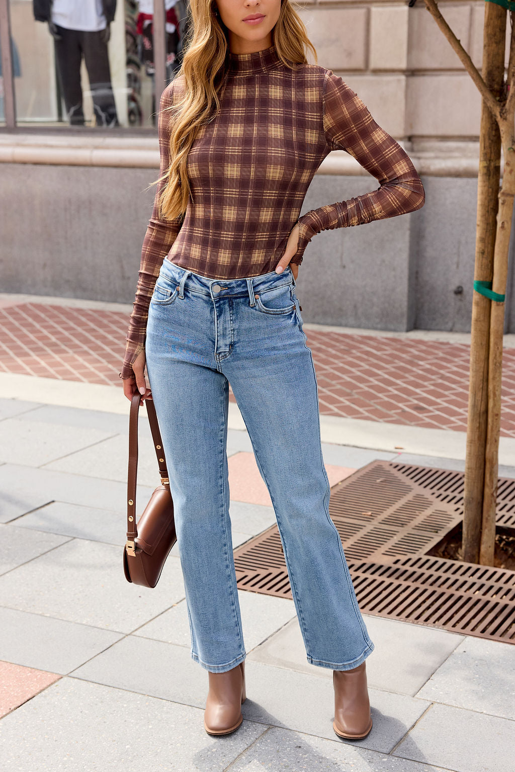 Woman in the Ruched Plaid Mesh Bodysuit - Brown, light blue jeans, and brown boots stands on a city sidewalk.