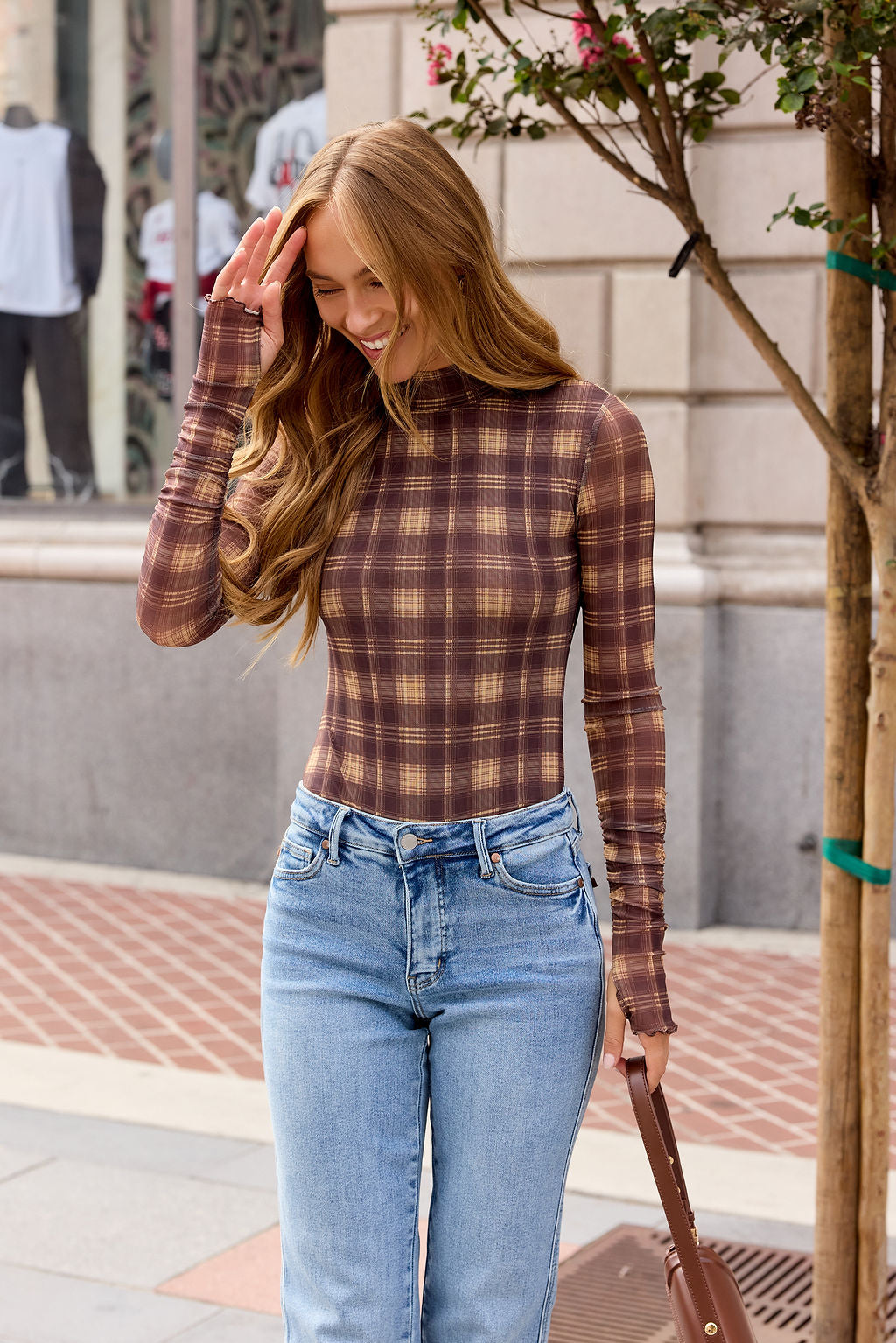 A woman in the Ruched Plaid Mesh Bodysuit - Brown and jeans smiles on a city sidewalk, holding a brown purse.