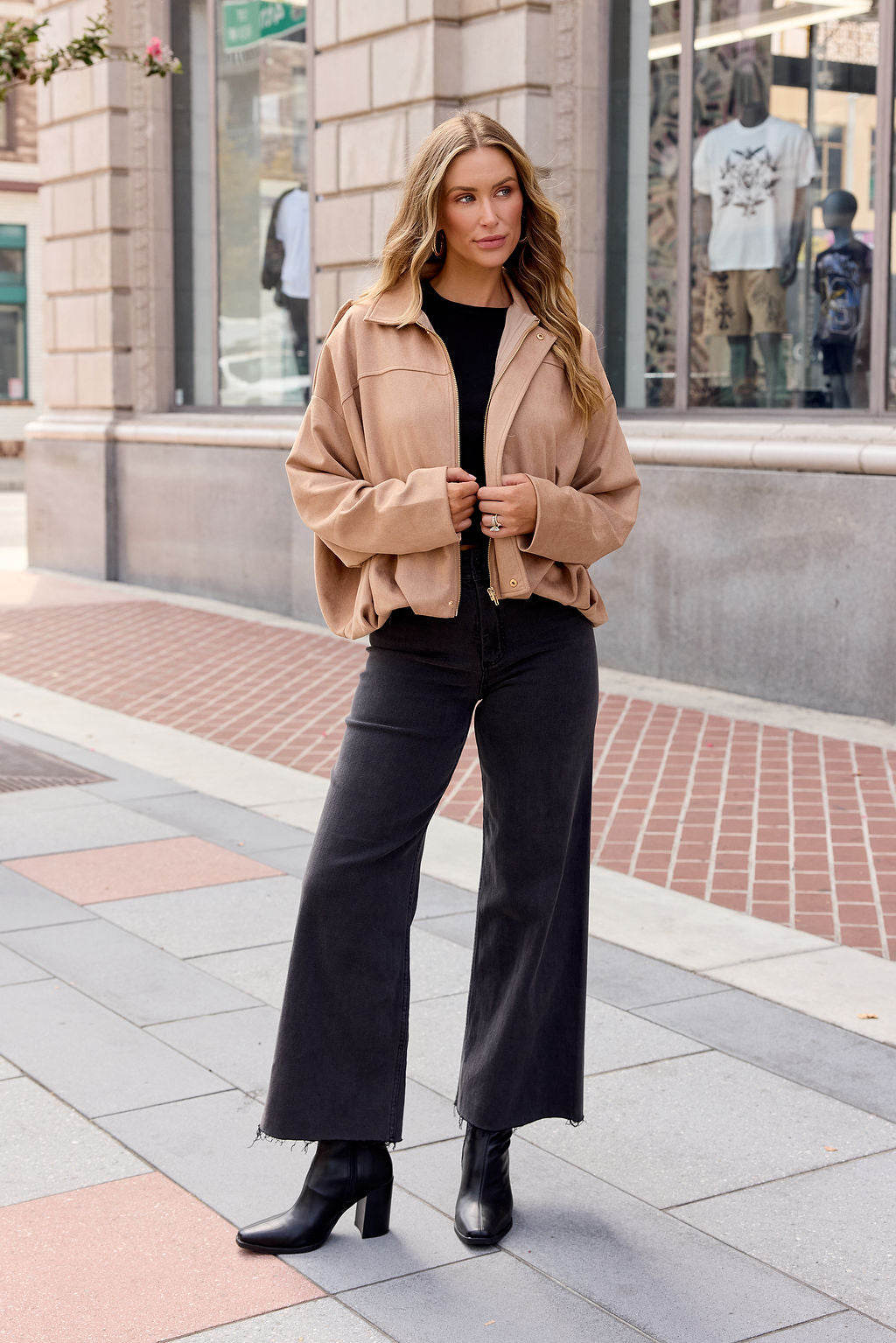 Woman in fall layers: Suede Bubble Hem Jacket - Camel, black top, wide jeans, boots on a city sidewalk near a shop window.