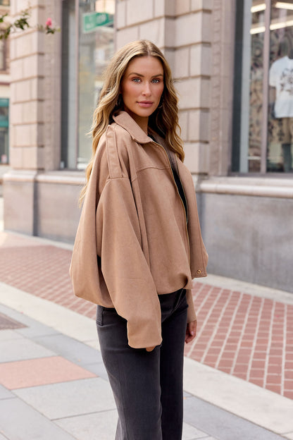 Woman with long wavy hair wears the Suede Bubble Hem Jacket - Camel and black pants, standing on a city sidewalk, looking at the camera.