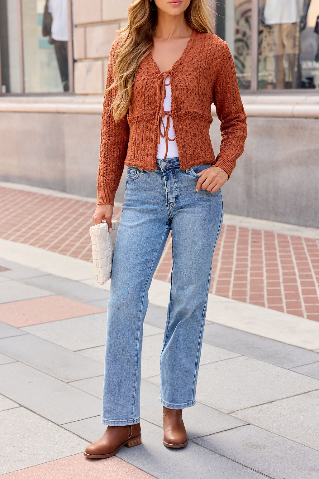 Woman wearing the Reese Tie Front Sweater - Terracotta, white top, blue jeans, brown boots, and holding a white woven clutch on sidewalk.
