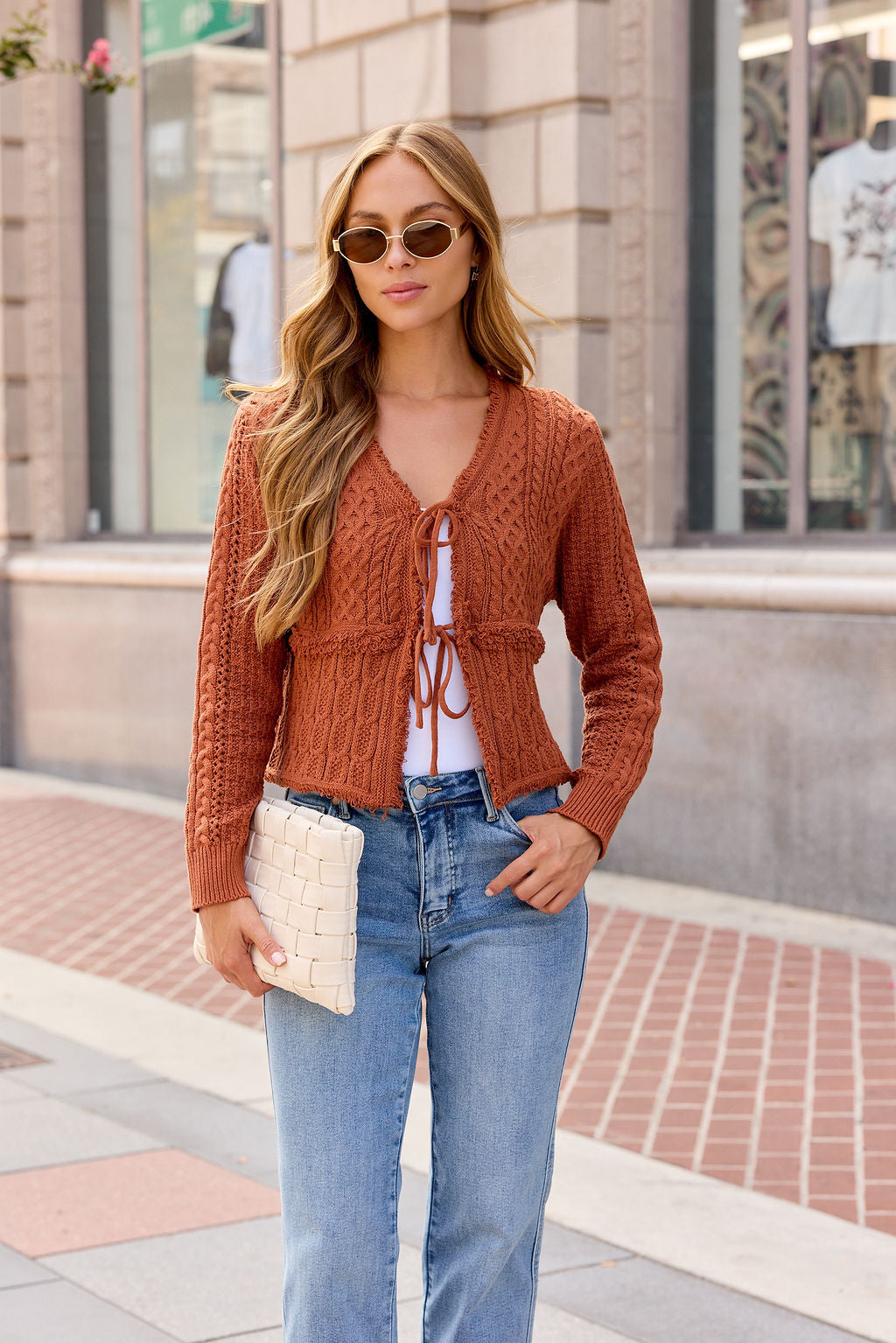 Woman in sunglasses wears the Reese Tie Front Sweater - Terracotta with jeans, holding a woven clutch on a city sidewalk.