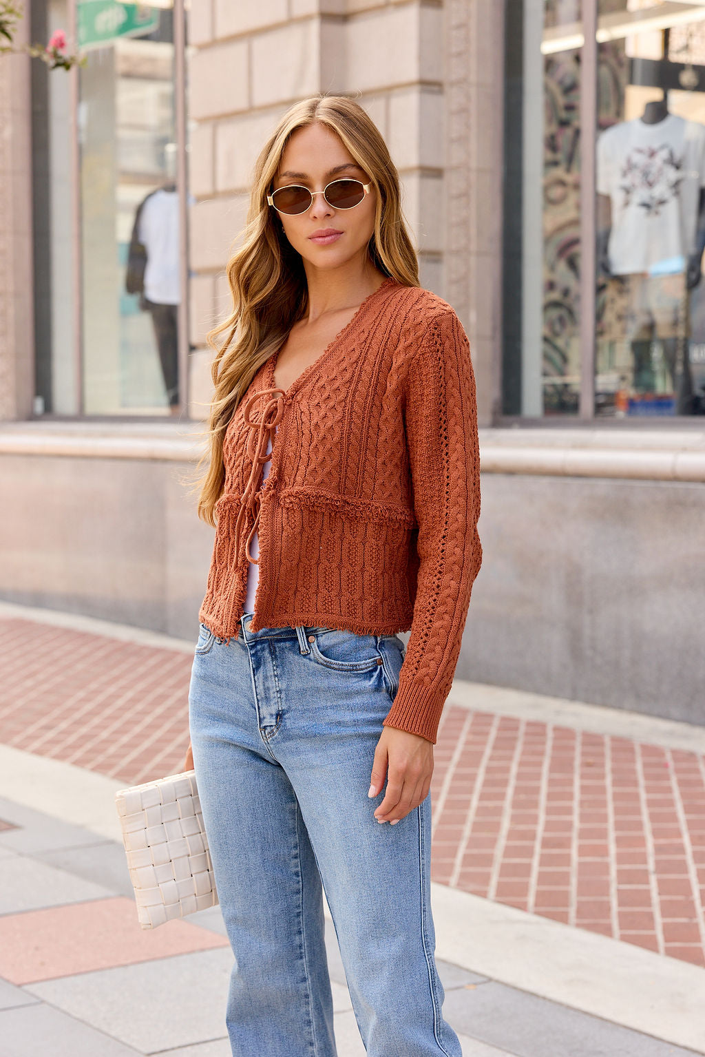 Woman with long hair in sunglasses and jeans wears the Reese Tie Front Sweater - Terracotta, holding a white clutch on a city sidewalk.