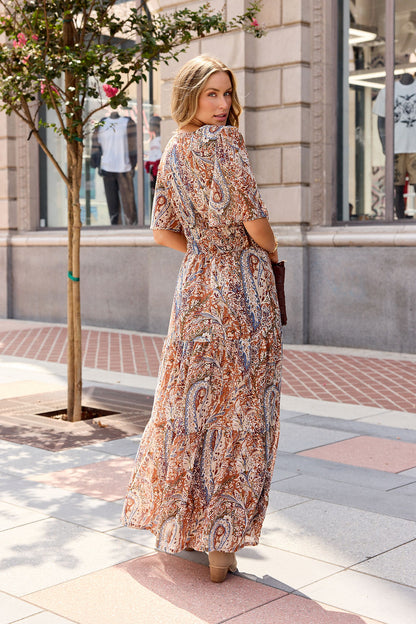 Woman in a Harvest Print Maxi Dress - Multi stands on a city sidewalk, glancing back over her shoulder near a tree.