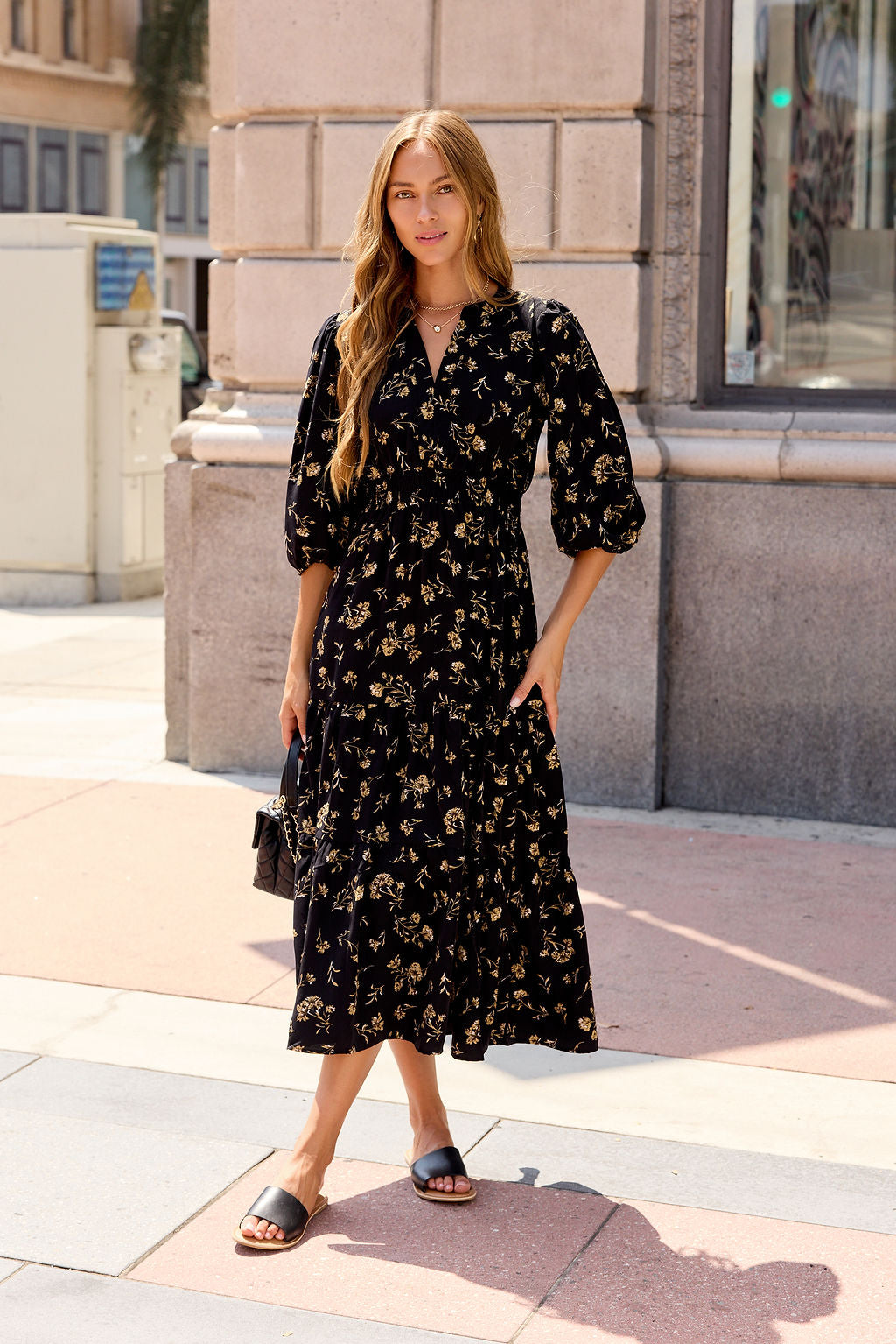 Woman in the Ember Floral Midi Dress - Black and sandals stands on a city sidewalk, holding a small purse.