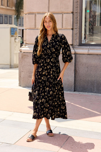 Woman in the Ember Floral Midi Dress - Black and sandals stands on a city sidewalk, holding a small purse.