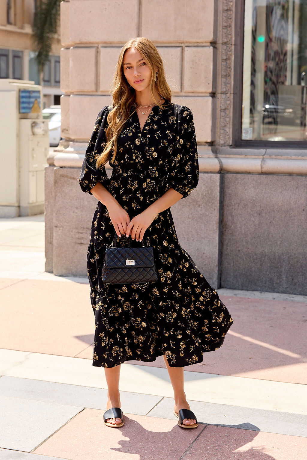Woman in the Ember Floral Midi Dress - Black, with a black purse and sandals, stands on a city sidewalk.