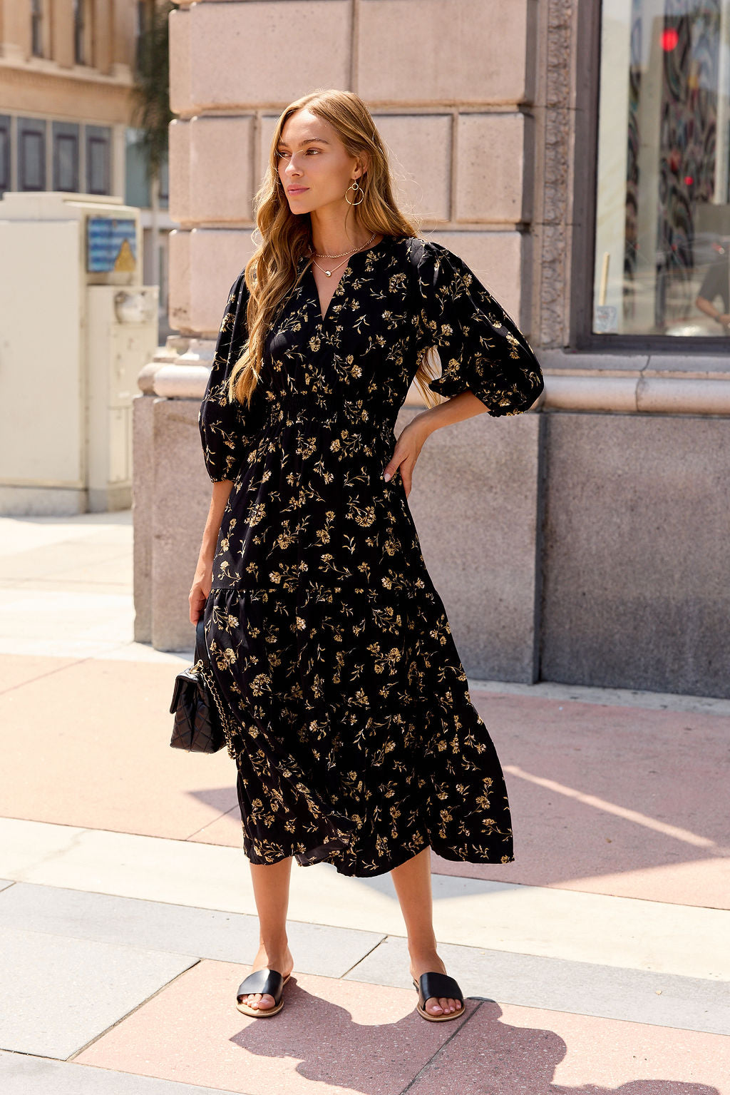 Woman in the Ember Floral Midi Dress - Black stands on a city sidewalk with sandals and a purse, sunlight and buildings behind her.