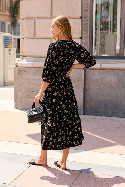 Woman in Ember Floral Midi Dress - Black with sandals holds a black quilted handbag on a sunny sidewalk.