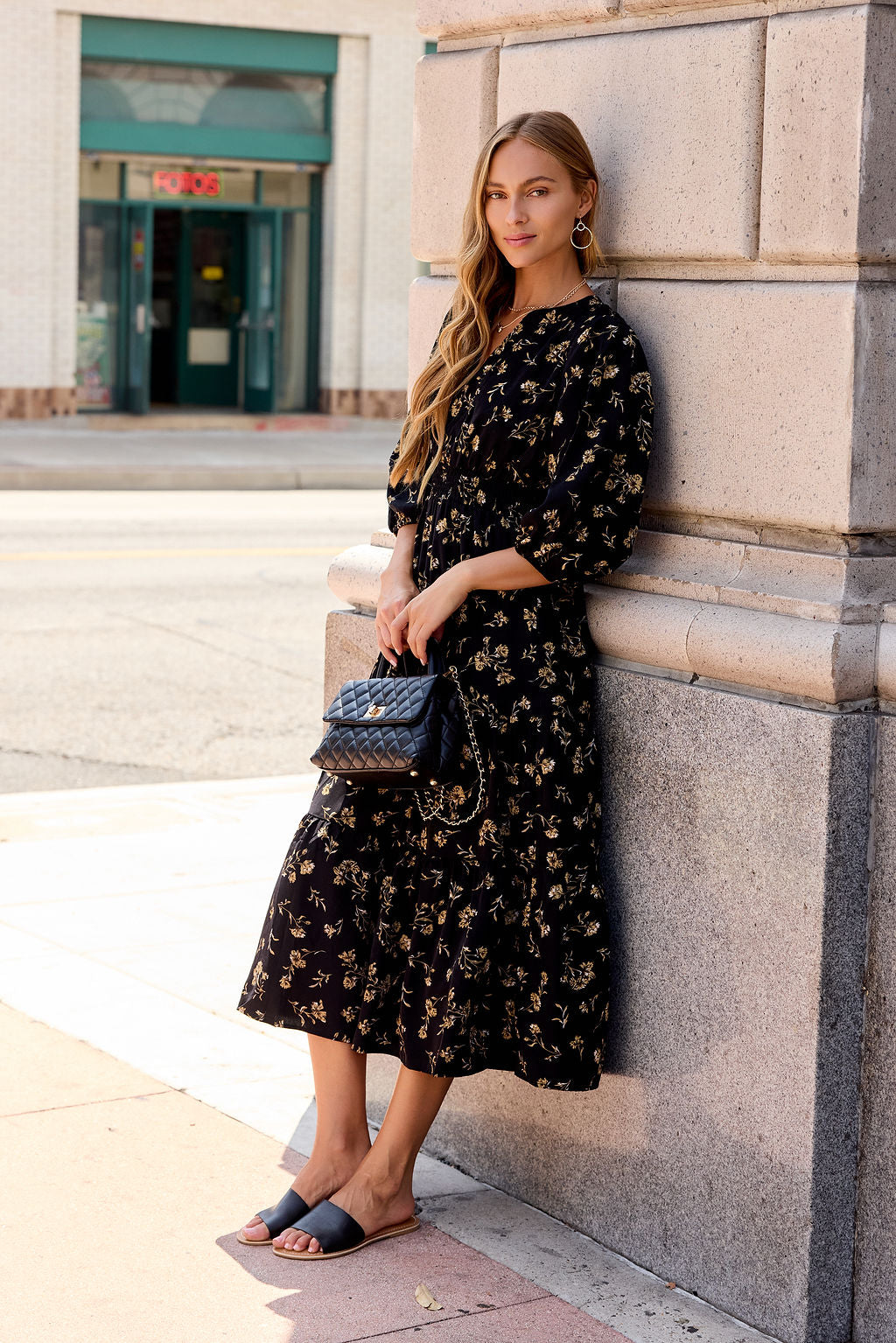 Woman wears Ember Floral Midi Dress - Black, leaning on a wall with a quilted purse in the sun on a city sidewalk.
