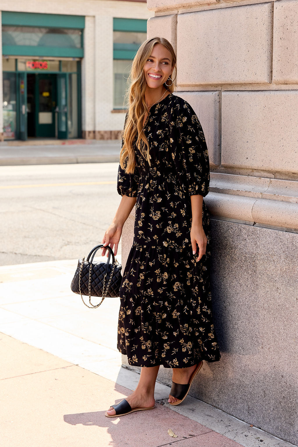 Woman in the Ember Floral Midi Dress - Black, sandals, and a black handbag smiles by a building.