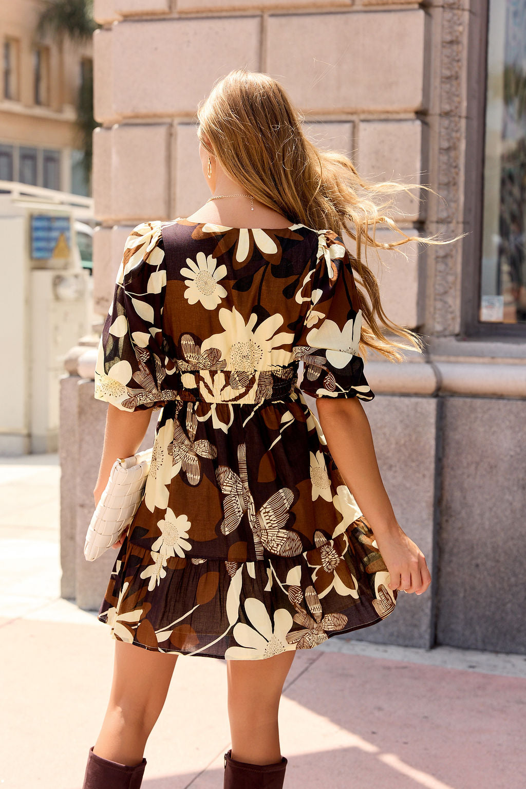 Woman with long hair in the Poppy Floral Mini Dress - Brown walks a sunny city street, wearing boots and holding a white clutch.