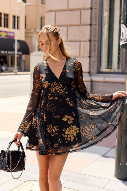 Woman in the Camellia Floral Mesh Mini Dress - Black stands on a city sidewalk, holding a black purse and smiling.