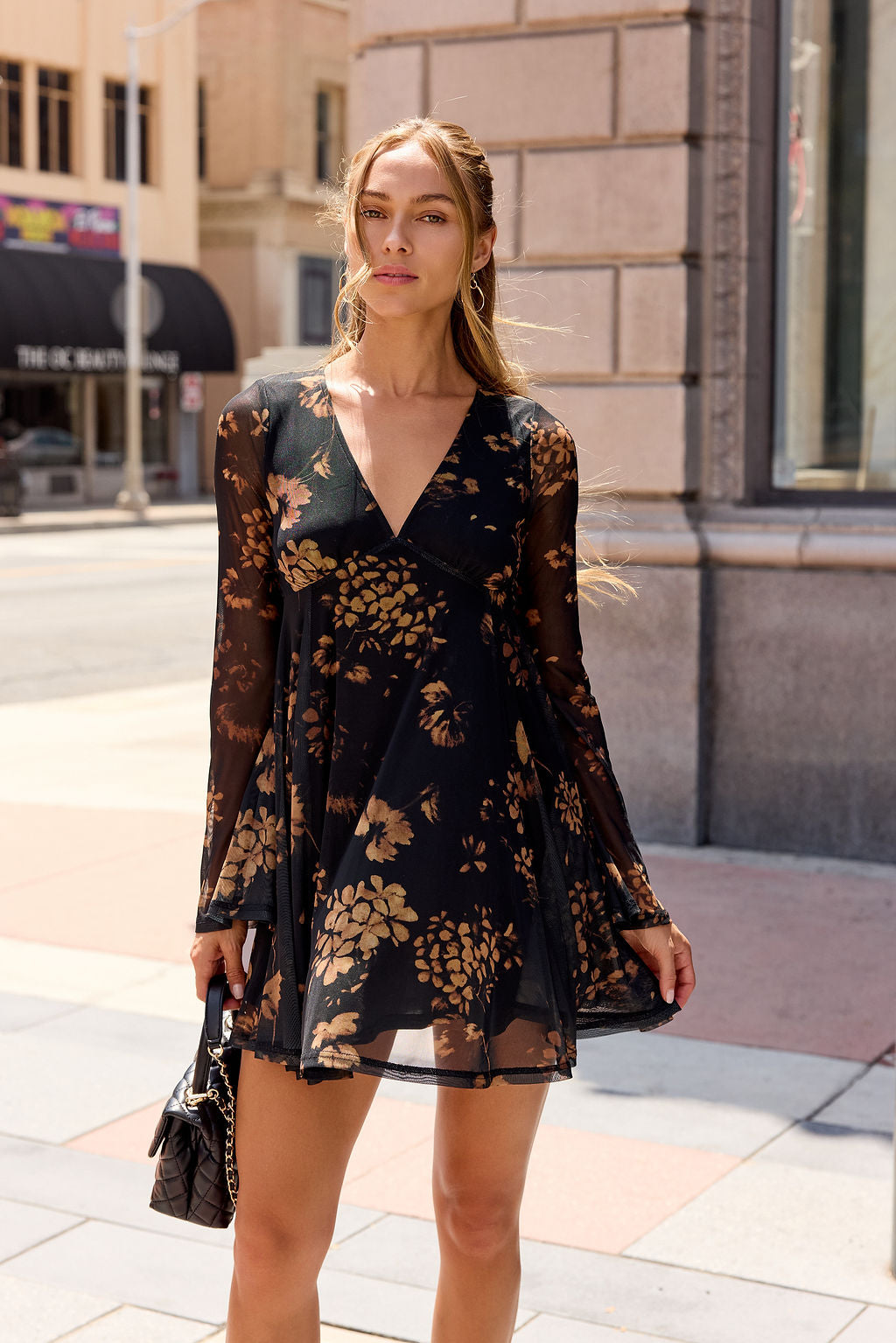 Woman in the Camellia Floral Mesh Mini Dress - Black stands on a city sidewalk, holding a black purse, looking confidently at the camera.
