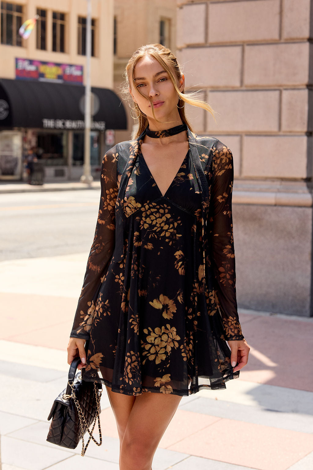 Woman in the Camellia Floral Mesh Mini Dress - Black with a choker, purse in hand, stands on a sunny city street—perfect date night look.