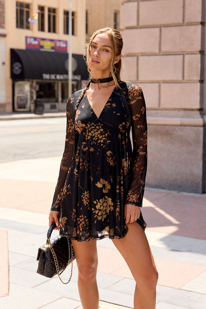 Woman in the Camellia Floral Mesh Mini Dress - Black, holding a black bag, stands on a sunny city sidewalk.