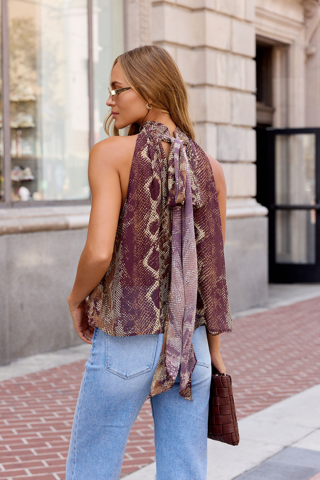 Woman with long hair in Steve Madden Alexane Top - Brown, light blue jeans, sunglasses, and holding a brown clutch on city sidewalk.
