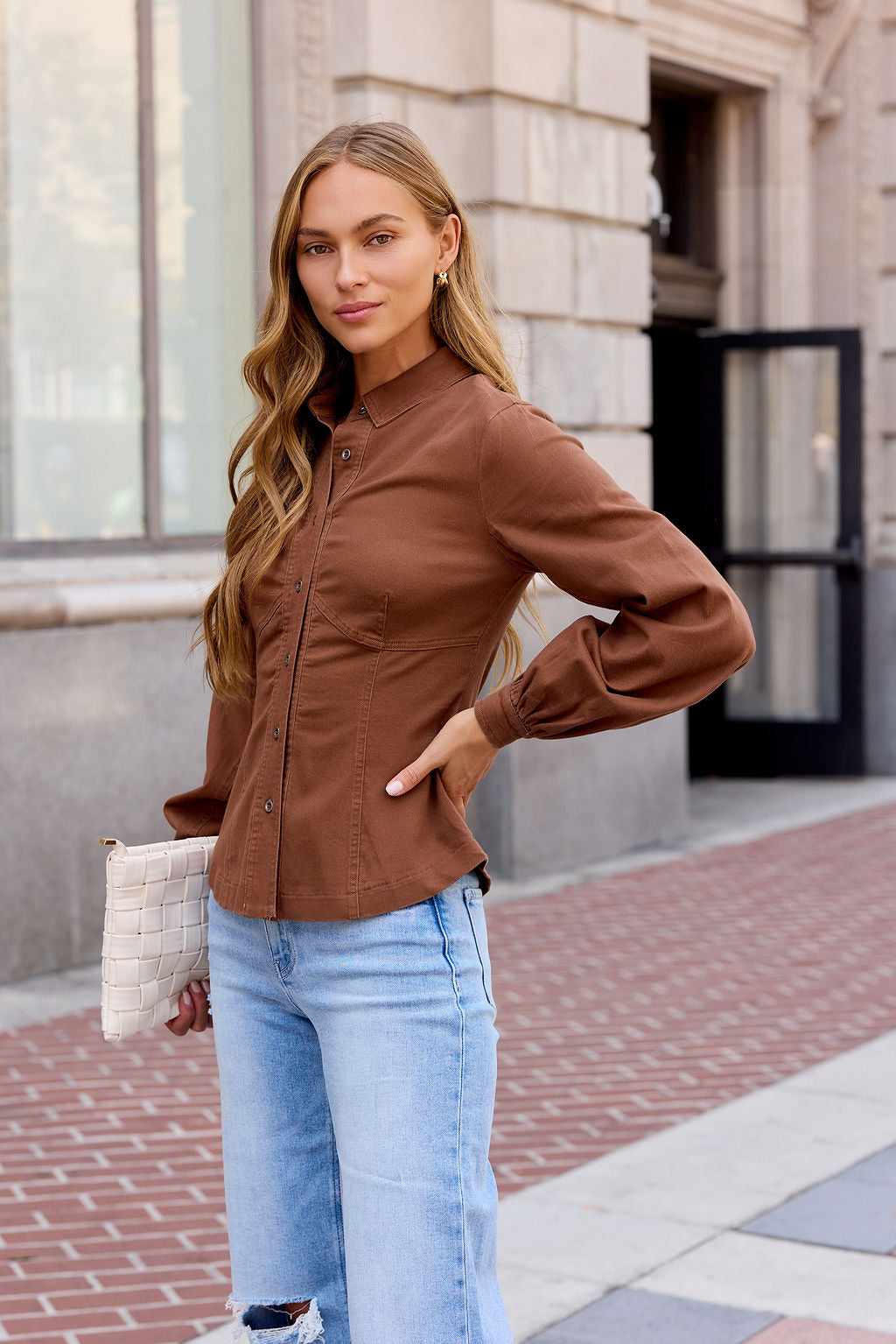 Woman in the Sutton Twill Button Down Top - Mocha and light jeans stands on a city sidewalk holding a white woven clutch.