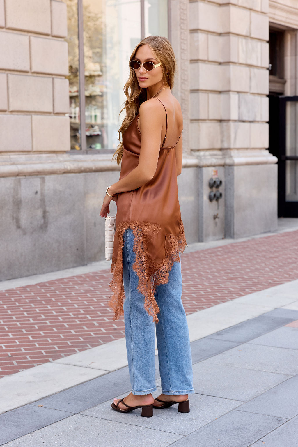 Woman in an Asymmetrical Satin Lace Top - Camel, blue jeans, and sandals stands on a city sidewalk, looking over her shoulder.