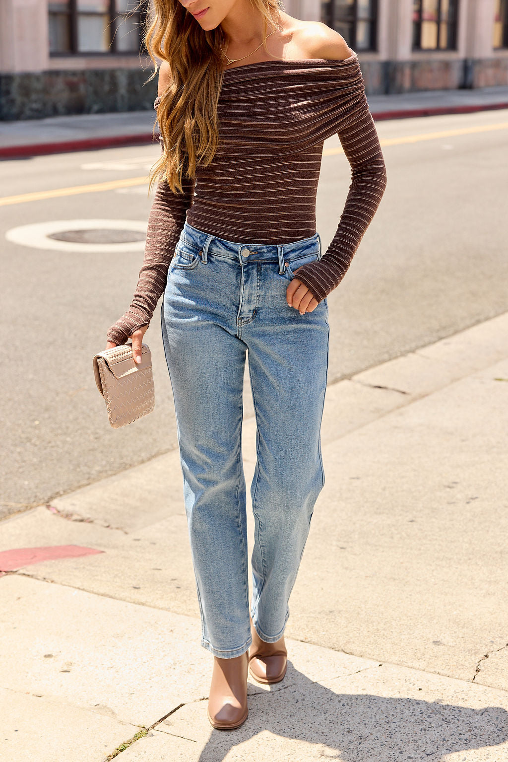 Woman in a Striped Off The Shoulder Bodysuit - Mocha, light blue jeans, and beige clutch poses stylishly on the sidewalk.