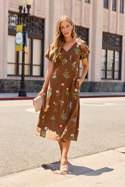 Woman wears the Wildflower Floral Midi Dress - Brown on a city sidewalk, holding a beige clutch with buildings and a streetlamp behind her.