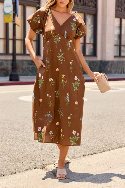 Woman in the Wildflower Floral Midi Dress - Brown holds a beige handbag, standing stylishly on a sunlit street.