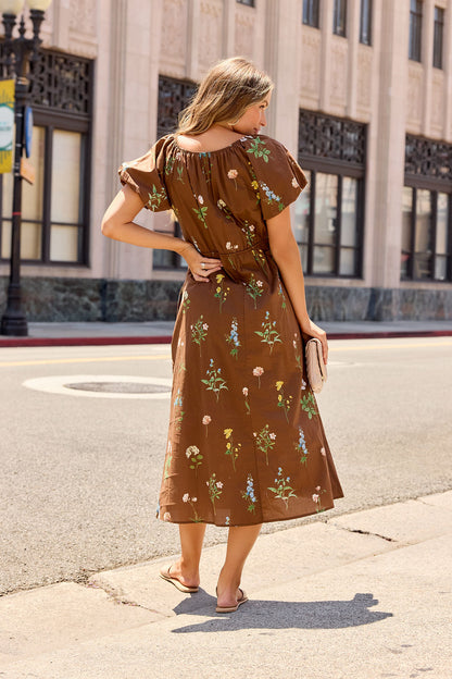 Woman in the Wildflower Floral Midi Dress - Brown and sandals, holding a beige clutch on a city sidewalk, faces away from the camera.